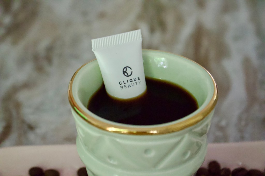 A small packet of Clique Beauty eye cream inserted in a decorative cup filled with dark liquid, surrounded by coffee beans.