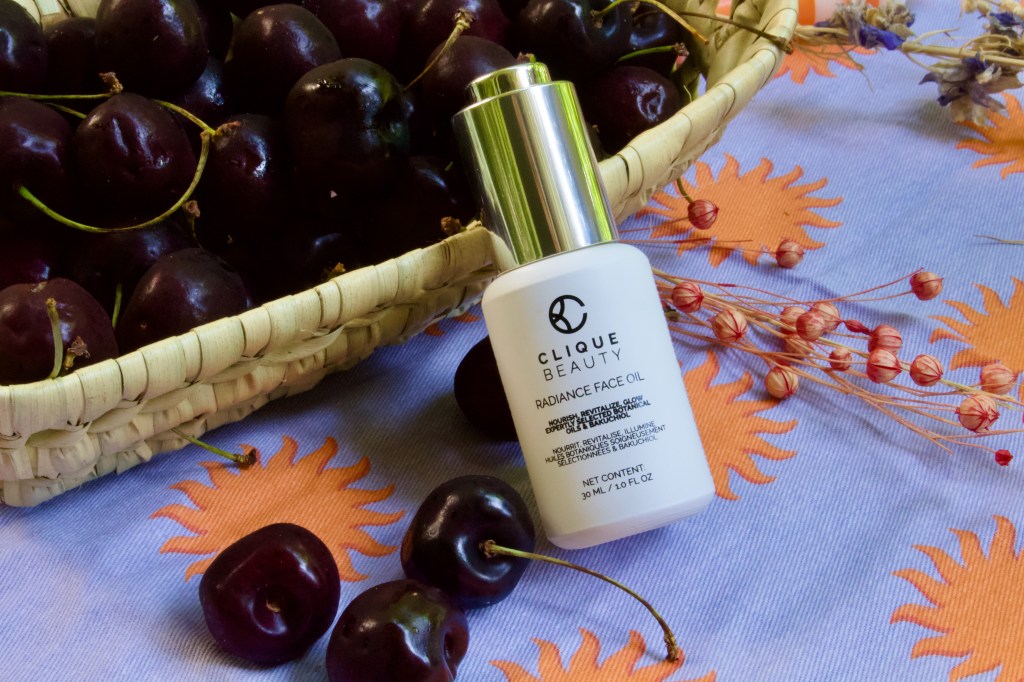 A bottle of Clique Beauty Radiance Face Oil placed next to a woven basket filled with dark cherries, surrounded by colorful floral decorations on a bright background.