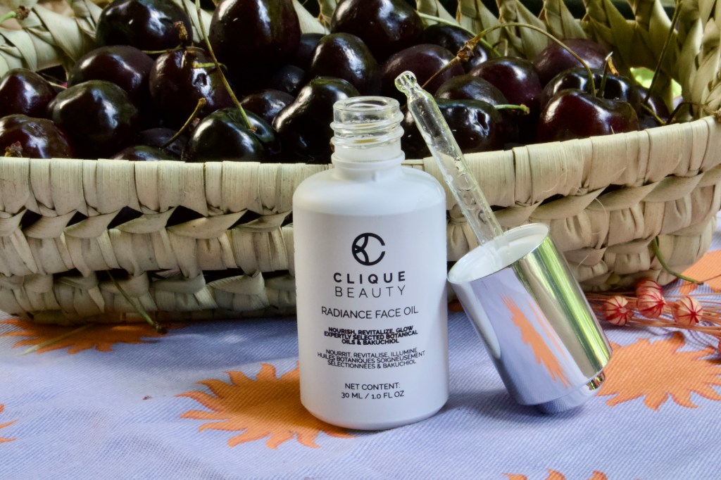 A bottle of Clique Beauty Radiance Face Oil with a dropper, placed next to a basket of dark cherries on a patterned tablecloth.